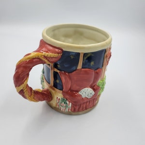 Pode incluir: Uma caneca de cer&acirc;mica com tema natalino com uma al&ccedil;a vermelha e um design detalhado. A caneca apresenta uma cena aconchegante da sala de estar com uma poltrona vermelha, um gato dormindo e uma janela com vista para uma noite estrelada.