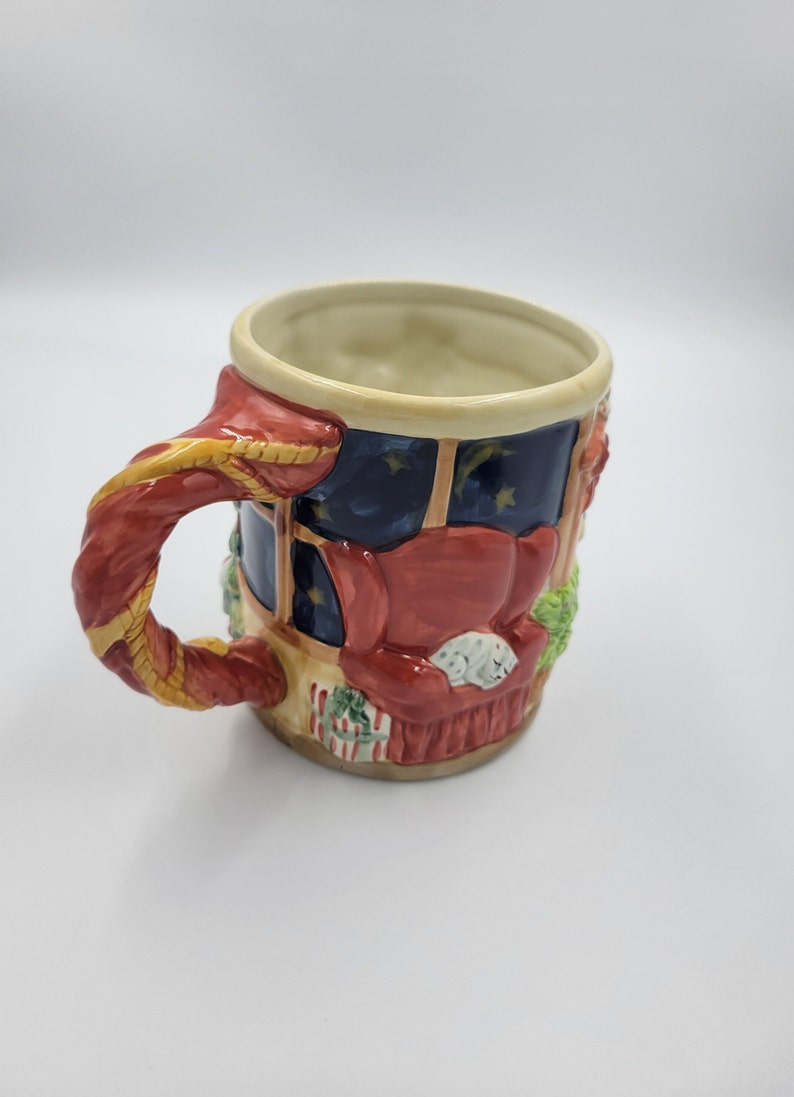 Pode incluir: Uma caneca de cer&acirc;mica com tema natalino com uma al&ccedil;a vermelha e um design detalhado. A caneca apresenta uma cena aconchegante da sala de estar com uma poltrona vermelha, um gato dormindo e uma janela com vista para uma noite estrelada.