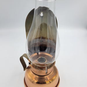 May include: A wall-mounted oil lamp featuring a polished copper base and a clear glass chimney. The lamp includes a curved metal handle on the side and a dark, rounded metal backplate with a keyhole slot for mounting on a vertical surface.