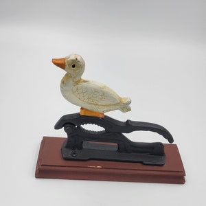 May include: A white duck nutcracker with orange accents sits on a black metal base with a wooden platform. The duck is perched on top of the nutcracker, which has a lever on the side.
