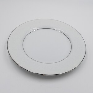 May include: White ceramic plate with a silver rim. The plate features a delicate, embossed pattern around the edge. The center of the plate is smooth and white. This elegant plate is suitable for formal dining.