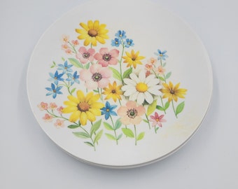 May include: White ceramic plate with a floral design featuring yellow, pink, blue, and white flowers. The flowers are arranged in a bouquet style with green leaves. The plate is round and has a simple, elegant design.