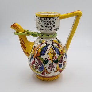 May include: A hand-painted ceramic coffee pot with a yellow handle and spout. The pot is decorated with colourful birds and floral patterns. Text on the pot reads "LE DIABLE L'ENFER UN ANGE L'AMOUR d pour le café."