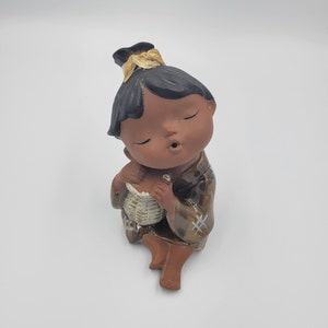 May include: A ceramic figurine of a seated child with closed eyes, holding a small woven basket. The child has dark hair with a gold accent and wears a brown kimono with a geometric pattern. The figurine is on a white background.