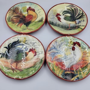 May include: Four decorative ceramic plates featuring rooster illustrations. Each plate has a different rooster design with vibrant colours, set against a background of script and a red rim. Suitable for kitchen or dining decor.