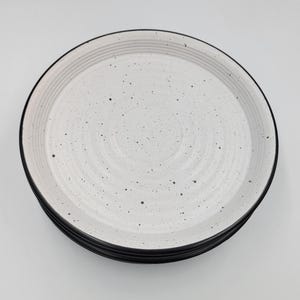 May include: A stack of speckled white ceramic plates with a black rim. The plates have a textured surface with concentric circles and small black speckles. The plates are stacked on top of each other.
