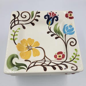May include: Square ceramic plate with a cream-colored background. The plate is decorated with colorful floral designs in yellow, blue, red, and green. Brown vines and swirls frame the flowers.