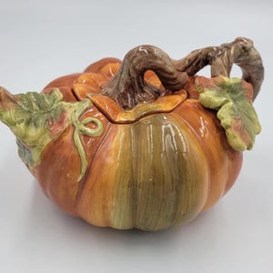 May include: A ceramic teapot shaped like a pumpkin with a glossy orange and green finish. The lid features a brown, textured stem handle, while the body is decorated with sculpted green leaves and vines on the sides, creating a realistic look.