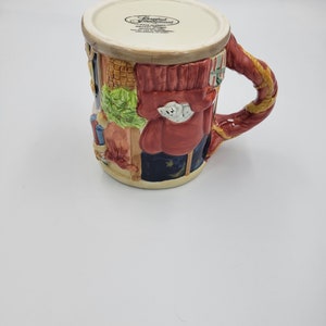 Pode incluir: Uma caneca de cer&acirc;mica com um design detalhado e colorido. A caneca apresenta um casaco vermelho, uma roupa azul e uma al&ccedil;a decorativa. A caneca tem uma borda creme e uma cena detalhada de uma pessoa. A caneca &eacute; um item decorativo.