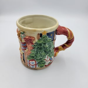 Pode incluir: Uma caneca de Natal de cer&acirc;mica com um design festivo. A caneca apresenta uma &aacute;rvore de Natal verde, presentes embrulhados e uma al&ccedil;a vermelha e dourada. O interior da caneca &eacute; creme e tem um exterior esculpido.