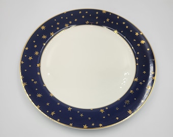 May include: A round, white ceramic plate with a navy blue border. The border is decorated with gold stars. A thin gold band lines the inner and outer edges of the navy blue border. The plate has a glossy finish.