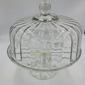 Peut inclure: Un présentoir à gâteau en verre transparent avec un couvercle bombé assorti. Le couvercle présente des détails nervurés verticaux et une poignée sphérique sur le dessus. La base est circulaire et surélevée, offrant un support stable.