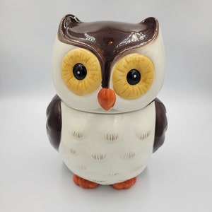 May include: A ceramic owl-shaped cookie jar. The jar features a brown top, white face, and brown wings. The eyes are large and black with yellow accents. The beak is orange, and the feet are orange. The jar is designed to look like a cartoon owl.