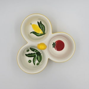 May include: A three-section ceramic serving dish with a yellow border. Each section features a different hand-painted vegetable: corn, peas, and a tomato. The dish has a yellow handle and is ideal for serving snacks or dips.