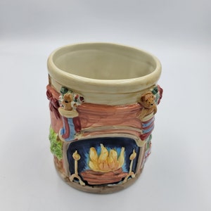 Pode incluir: Uma caneca de cer&acirc;mica com tema natalino com borda e interior creme. O exterior apresenta um design de lareira com fogo ardente, meias e um ursinho de pel&uacute;cia. A caneca &eacute; pintada em tons de vermelho, azul, verde e castanho.