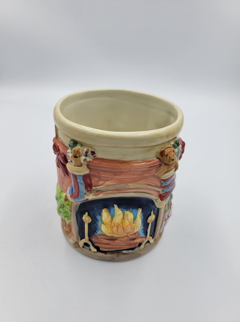 Pode incluir: Uma caneca de cer&acirc;mica com tema natalino com borda e interior creme. O exterior apresenta um design de lareira com fogo ardente, meias e um ursinho de pel&uacute;cia. A caneca &eacute; pintada em tons de vermelho, azul, verde e castanho.