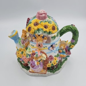 May include: A whimsical ceramic teapot featuring a house design with bunnies, sunflowers, and butterflies. The teapot has a green handle and a blue spout. The lid is topped with a pink rose. The base is white with colorful accents.