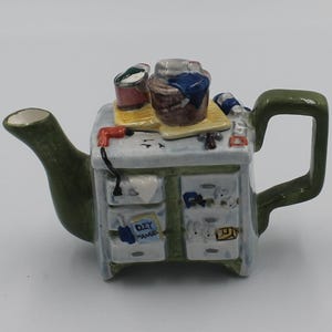 May include: A ceramic teapot shaped like a workbench with a green spout and handle. The top surface features miniature tools and a bucket. The front displays drawers, a blue book labeled DIY MANUAL, and various small items painted in detail on the surface.