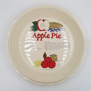 May include: A white ceramic pie plate with a red apple and a pie crust design. The plate has a scalloped edge and features a recipe for apple pie with ingredients and instructions. The text on the plate reads "Apple Pie" and includes the copyright information "© LTD. COMMUNITIES, INC. 1982".