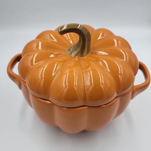May include: A glossy orange ceramic pumpkin-shaped lidded casserole dish with a brown stem handle on top. The round pot features deep vertical ridges and two curved side handles, providing a functional and decorative kitchen serving vessel.