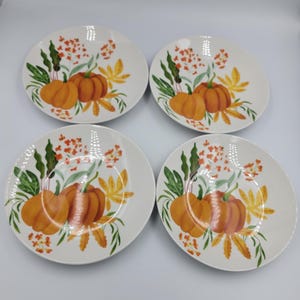 May include: A set of four round white ceramic plates, each featuring an identical illustration of orange pumpkins, green leaves, and small orange floral clusters arranged in a decorative pattern across the surface of the dinnerware.