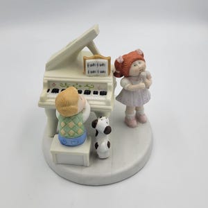 May include: A porcelain figurine set featuring a child playing a white grand piano with a music book, a child with red hair singing into a microphone, and a small black and white dog, all mounted on a circular base with a smooth white finish.