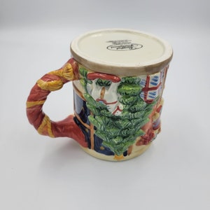 Pode incluir: Uma caneca de Natal de cer&acirc;mica com um design festivo. A caneca apresenta um relevo detalhado de uma &aacute;rvore de Natal, um boneco de neve e uma janela com um padr&atilde;o vermelho e azul. A al&ccedil;a &eacute; vermelha e dourada. Fundo branco.