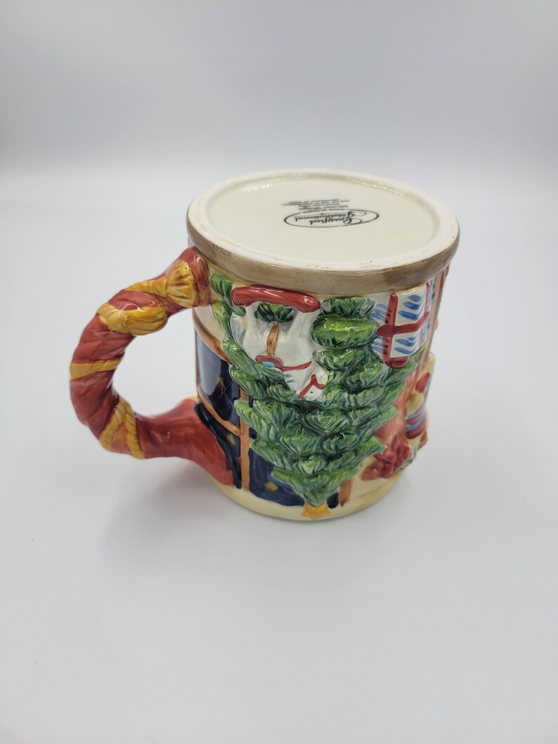 Pode incluir: Uma caneca de Natal de cer&acirc;mica com um design festivo. A caneca apresenta um relevo detalhado de uma &aacute;rvore de Natal, um boneco de neve e uma janela com um padr&atilde;o vermelho e azul. A al&ccedil;a &eacute; vermelha e dourada. Fundo branco.