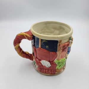 Pode incluir: Caneca de cer&acirc;mica decorativa com um design detalhado. A caneca apresenta uma al&ccedil;a vermelha com detalhes dourados e retrata uma sala com um sof&aacute; vermelho, um gato dormindo e uma janela com um c&eacute;u estrelado. Um item decorativo.