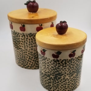 May include: Two ceramic canisters with wooden lids and apple-shaped knobs. The canisters are decorated with a black and white leopard print pattern and a band of red apples with green leaves around the middle.