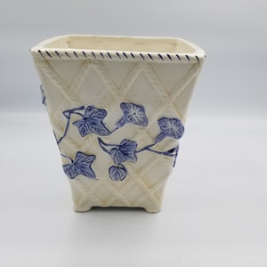 May include: Cream-colored square ceramic planter with a diamond pattern and blue floral accents. The planter has a woven basket design with blue ivy and flower details. The rim is trimmed with a blue rope design.