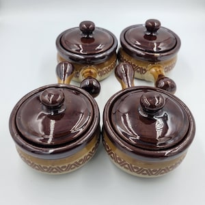 May include: A set of four round ceramic soup crocks with lids and side handles. Each crock features a dark brown glazed lid and rim, with a tan base decorated by a repeating geometric brown pattern around the exterior circumference.