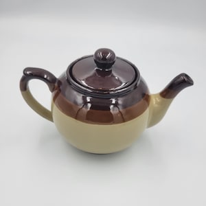 May include: A ceramic teapot with a rounded body and a lid. The teapot is primarily beige with a brown band around the middle and a dark brown top and handle. The spout and handle are curved.