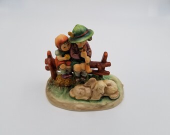 May include: A ceramic figurine depicting two children sitting on a wooden fence, one playing a flute. The figurine features a green hat, brown clothing, and a small lamb. The base is oval-shaped with grass and a brown fence.