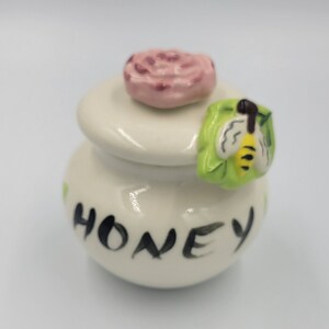 Peut inclure: Petit pot à miel en céramique blanche avec couvercle, orné d'une rose rose sur le dessus et d'une poignée verte en forme d'abeille. Le mot "HONEY" est écrit en script noir sur le devant. Le pot est de style fantaisiste et décoratif.