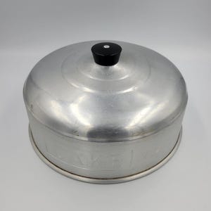 May include: A vintage aluminum cake carrier with a domed lid and a black knob. The carrier has a circular design and a silver finish. The word "CAKE" is partially visible on the side.