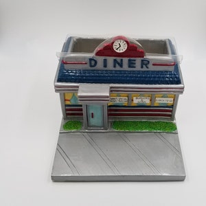 May include: A ceramic diner-shaped pencil holder with a red roof, blue and white striped walls, and a clock on the front. The holder has a sign that reads "Diner" and has signs that say "Today's Special -  Whims Meals", "Burgers & Soda", and "Lunch to Go".