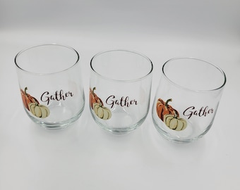 May include: Three clear stemless wine glasses with a fall theme. Each glass features a watercolour illustration of a pumpkin and the word "Gather" in a dark brown script. The pumpkins are orange and cream coloured.
