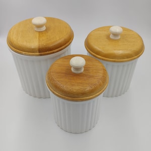 May include: Three white ceramic canisters with ribbed sides and wooden lids. Each lid has a small, round, off-white knob. The canisters are of varying sizes, creating a tiered arrangement. The lids are a light brown color.
