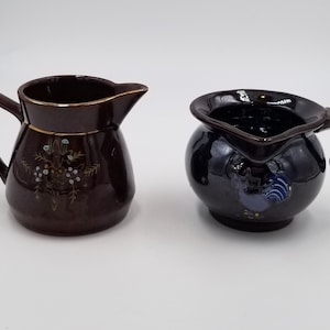 May include: Two ceramic pitchers. One is brown with a gold rim and floral design. The other is dark blue with a rounded shape and a rooster design. Both have handles and spouts.