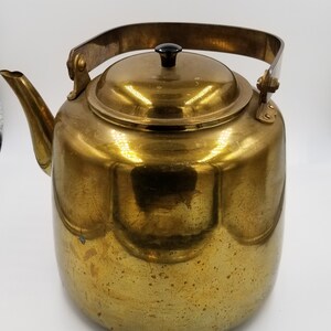 May include: A vintage brass kettle with a curved spout, a hinged lid, and a black knob. The kettle has a large handle and is designed for stovetop use. The brass has a warm, golden color and shows signs of age.