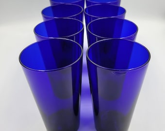May include: A set of eight cobalt blue drinking glasses arranged in four rows of two. Each glass features a smooth, tapered cylindrical shape with a wide circular rim and a solid base, showcasing a consistent deep blue translucent glass material.
