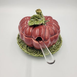 May include: A ceramic pumpkin-shaped serving dish with a lid and a spoon. The dish is a deep red color, with green leaves and a stem on top. It sits on a green leaf-shaped plate.