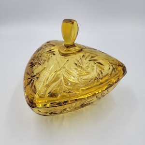 May include: Amber-colored, heart-shaped glass trinket dish with a lid. The dish features a detailed, embossed floral pattern and a small, rectangular handle on the lid. The glass has a glossy finish, reflecting light. A decorative storage container.