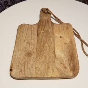 May include: A rectangular wooden cutting board with a handle and a rope for hanging. The board is made of light brown wood with visible grain and knots. The rope is a natural tan color. The board is designed for kitchen use.