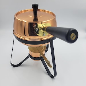 May include: Copper fondue pot with a black handle and a black metal stand. The pot has a lid with a black knob.