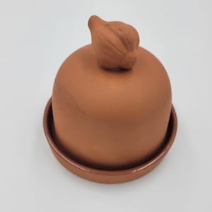 May include: A terracotta butter bell with a smooth, matte finish. The dome-shaped lid features a sculpted handle shaped like a garlic bulb at the top. The base is a circular, shallow dish that holds the dome, both in a consistent reddish-brown clay.