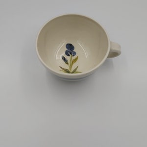 May include: Cream-colored ceramic mug with a blue floral design inside. The mug has a handle and a blue stripe around the base. The floral design features a blue flower with green leaves.