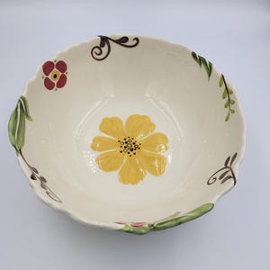 May include: Cream-colored ceramic bowl with a scalloped edge. The bowl features a large yellow flower in the center and smaller floral and leaf designs around the rim. The bowl is hand-painted with red, green, and brown accents.