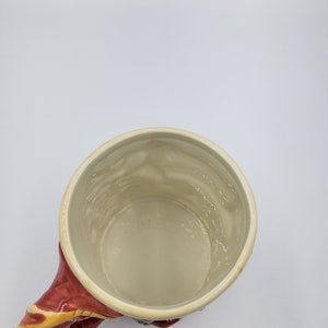 Pode incluir: Uma caneca de cer&acirc;mica com interior creme e uma al&ccedil;a vermelha com detalhes dourados e verdes. O interior da caneca tem uma superf&iacute;cie texturizada. A caneca est&aacute; sobre um fundo branco.